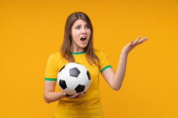 Young female soccer fan or player holding a soccer ball on yellow background, looking aside with a judging, disappointed and frustrated expression