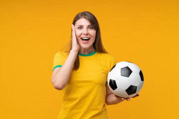 Surprised young woman in yellow shirt holding soccer ball and touching her face in amazement