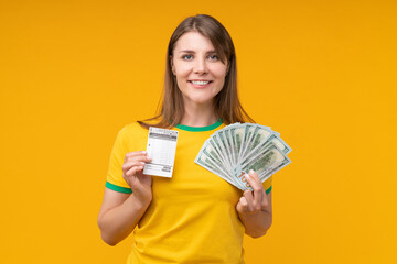 Young woman posing on yellow background holding a betting slip ticket in one hand and a fan of dollar bills in the other, smiling with joy