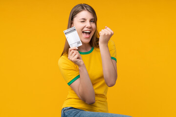Young woman on yellow studio background holding a lottery ticket, winking and making a yes gesture...