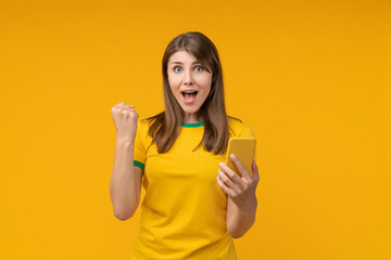 Young woman on yellow studio background holding a smartphone in one hand and making a yes fist gesture with an overjoyed expression, celebrating a big win in betting, gambling, and victory
