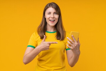Young woman on yellow studio background winking and pointing at a smartphone in her hand, symbolizing confidence and excitement in betting, gambling, and winning with bookmaker apps