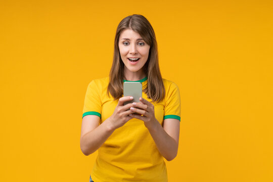 Young woman on yellow studio background reading a message on her smartphone with a surprised expression, reacting to unexpected betting results, gambling win, or bookmaker app notification