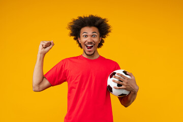 Happy African man on yellow background holds a soccer ball in one hand and makes a yes gesture with...