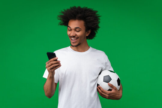 Joyful young African man holding a soccer ball and looking at his smartphone screen