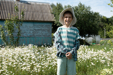 a boy in the countryside is standing in front of a village house and smiling, next to him after the daisies