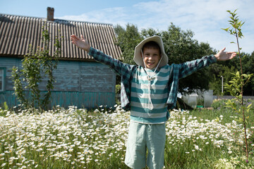 a young boy in the countryside is standing in front of a village house, his arms raised and smiling