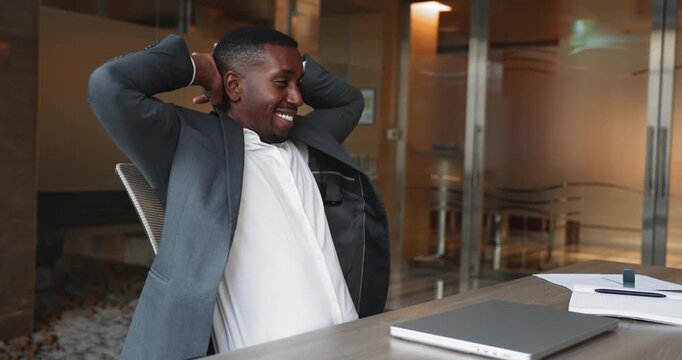 Happy successful African American businessman finishing work day, completing project task, closing computer, placing hands on nap, leaning on chair back, relaxing with toothy smile