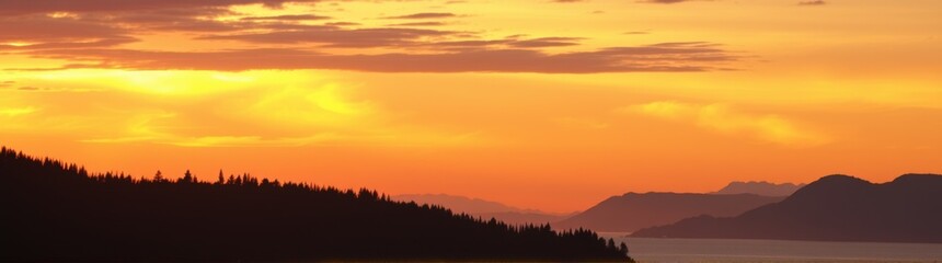 Fototapeta premium Mountains and trees are silhouetted against a sunset sky with a boat in the water banner