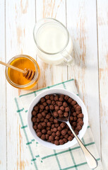 chocolate corn balls in a ceramic bowl with milk and honey on a white wooden table, top view.
