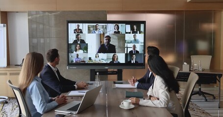 Diverse team of four young business professional colleagues meeting on online hybrid conference, sitting at shared office table, looking at wall screen with remote coworkers on video call - Powered by Adobe