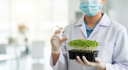 A scientist in a lab coat and mask holds a tray of seedlings and a syringe