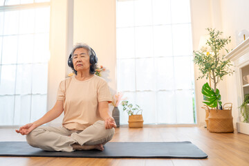 Happy Asian senior woman meditating in lotus pose with sound therapy on headphones at home. Healthy elderly woman enjoy wellness healthcare lifestyle relaxing, meditation yoga exercise in living room.