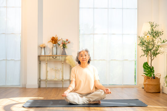 Happy Asian senior woman sit on yoga mat, meditating in lotus pose at home. Healthy elderly woman enjoy wellness healthcare lifestyle relaxing, meditation deep breath and yoga exercise in living room.