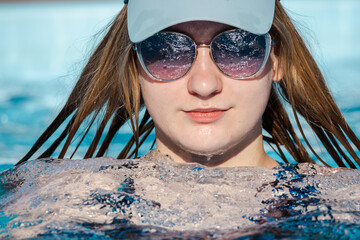 Teenage girl wearing sunglasses and a cap relaxing in a swimming pool, with wet hair and calm...