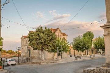 In the fading light of day, a charming church stands in a tranquil Greek village. Benches line the...