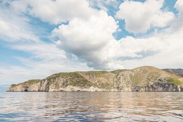 Tranquil waters meet a dramatic coastline of rocky cliffs in Greece. Lush vegetation covers the...
