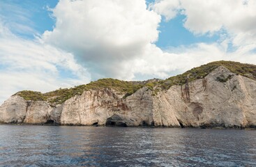 A boat glides along the coast revealing towering cliffs riddled with caves. Lush green vegetation...