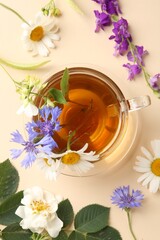 Aromatic herbal tea in glass cup and different flowers on beige background, flat lay