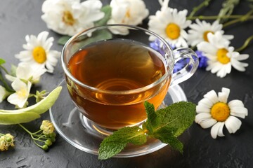 Aromatic herbal tea in glass cup, different flowers and mint on black table, closeup