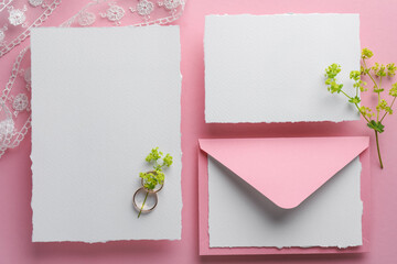 Wedding invitation cards, envelope, golden rings, lace ribbon and branches with flowers on pink background, flat lay