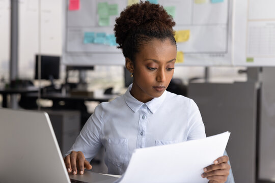 African woman sits at desk summarizing information from laptop onto paper, planning tasks based on document, researching, comparing data, looks focused. Workflow using printed materials and internet