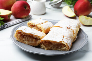 Pieces of tasty apple strudel with powdered sugar and fruits on white marble table, closeup