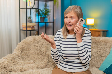 Caucasian mature woman sits on home sofa, arguing on phone, showing irritation. Grandmother deals...