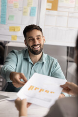 Smiling Latin businessman sits at desk handing document with bar charts and infographic data shown in report to client or colleague during negotiations, vertical shot. Workflow, statistics, analytics