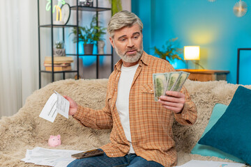 Mature man sitting on home sofa, using smartphone with calculator app to count money dollars cash....