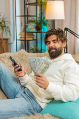 Planning budget. Rich happy Indian man using smartphone calculator app count dollar cash, calculate...
