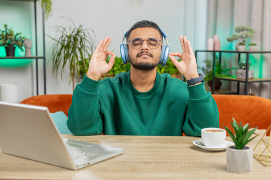 Keep calm down, relax. Indian man listening music breathes deeply, eyes closed meditating with concentrated thoughts, peaceful mind. Young bearded Arabian guy sits at home office room table workplace