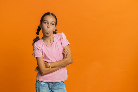 Cheerful funny young school girl showing tongue making faces at camera, fooling around, joking, aping with silly face teasing bullying abuse. Preteen female child kid 10 years old on orange background