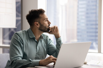 Pensive Latin man distracted from workflow looking out window to big city with contented smile,...