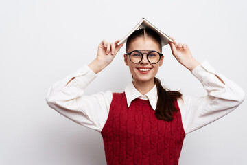 Smiling girl with book on head, playful pose in a white background, wearing a red sweater vest and...