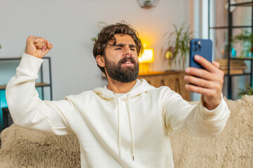 Happy young man using smartphone typing browsing saying yes, celebrating success victory, winning...
