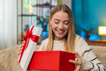 Happy Caucasian young woman girl sitting on couch at modern home opening birthday surprise box...