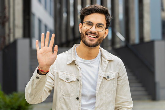 Hello. Indian man smiling friendly at camera, waving hands gesturing hi, greeting or goodbye welcoming with hospitable expression outdoors. Excited Arabian Hindu guy standing in urban city town street