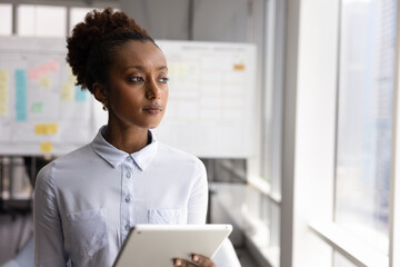 Young thoughtful African professional businesswoman stands alone by window holding tablet, gazing...