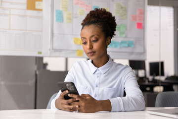 Serious African woman using smartphone seated in workplace, checking work emails or messages, chatting with colleague via messaging app, reviewing work calendar or schedule through mobile application