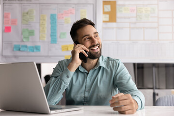 Smiling man lead talk on smartphone seated in workplace with open laptop, enjoy pleasant personal...