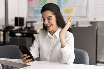 Happy businesswoman makes celebratory gesture while reading great news using her smartphone, sit at...