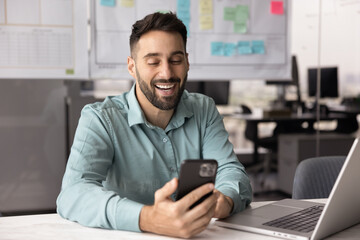 Happy Hispanic businessman sits at desk using his smartphone in modern workplace, reading good...