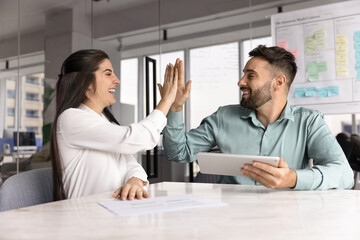 Two happy Latin teammates giving high five accomplished successful, productive teamwork using digital tablet seated at desk in modern meeting room. Partnership, innovations, professional achievements