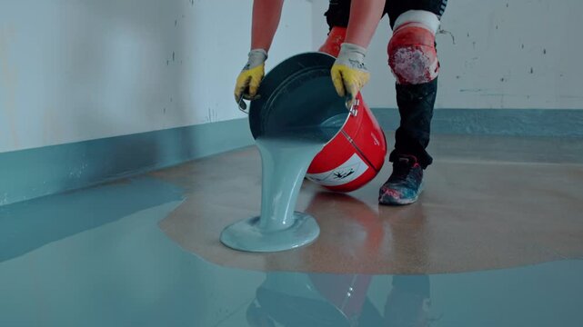 Worker pours light blue self-leveling epoxy resin from a red bucket onto a concrete floor
