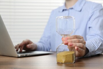 Deadline. Woman with hourglass and laptop working at wooden table in office, closeup