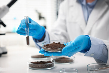 Scientist holding Petri dish with soil sample and test tube at white table in laboratory, closeup