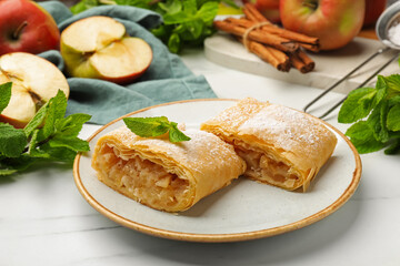 Pieces of tasty apple strudel with powdered sugar, mint, fruits and cinnamon on white marble table, closeup