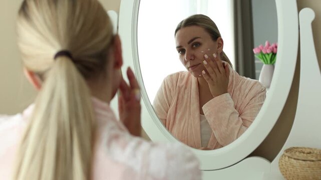 Woman applying cream onto face near mirror indoors. Acne treatment