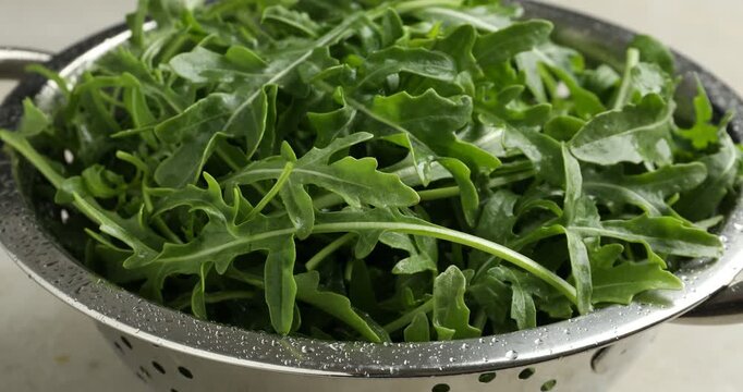 Fresh wet green arugula leaves in metal colander on white table, closeup. Camera zooming in